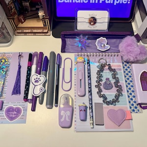 May include: A collection of purple stationery items including a daily planner, a small notebook with a gothic window sticker, various pens, markers, a pencil, a purple pencil case, a heart-shaped charm, a bow clip, and a beaded keychain.