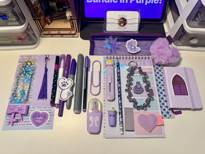 May include: A collection of purple stationery items including a daily planner, a small notebook with a gothic window sticker, various pens, markers, a pencil, a purple pencil case, a heart-shaped charm, a bow clip, and a beaded keychain.