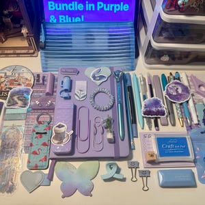 May include: A collection of stationery and craft supplies in shades of purple and blue, including a central purple notebook, various pens, a blue ink pad, flamingo-patterned items, butterfly-shaped paper, stickers, and metal binder clips.