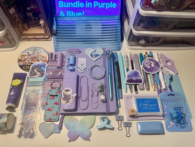 May include: A collection of stationery and craft supplies in shades of purple and blue, including a central purple notebook, various pens, a blue ink pad, flamingo-patterned items, butterfly-shaped paper, stickers, and metal binder clips.