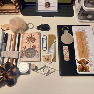 May include: A collection of stationery and reading accessories including a black notebook with a book tracker insert, a tarot card, a leopard print scrunchie, a beige pom-pom keychain, pens, a bear sticker, a bookmark with a white tassel, and a clear page holder.
