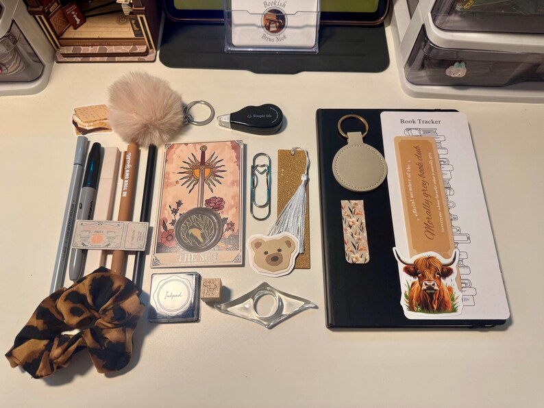 May include: A collection of stationery and reading accessories including a black notebook with a book tracker insert, a tarot card, a leopard print scrunchie, a beige pom-pom keychain, pens, a bear sticker, a bookmark with a white tassel, and a clear page holder.