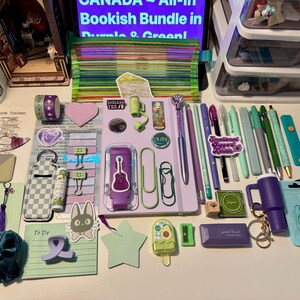 May include: A collection of stationery and accessories in shades of purple and green, including a notebook, pens, pencils, paper clips, a scrunchie, stickers, a small pouch, and various desk supplies arranged on a flat surface.