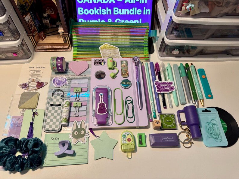 May include: A collection of stationery and accessories in shades of purple and green, including a notebook, pens, pencils, paper clips, a scrunchie, stickers, a small pouch, and various desk supplies arranged on a flat surface.