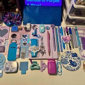 May include: A collection of stationery and accessories in shades of purple and blue, including a notebook, pens, a pencil, a scrunchie, stickers, a circular cat-themed coaster, a blue pencil case, clips, a hair claw, and various small desk items.
