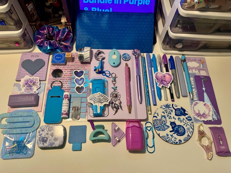 May include: A collection of stationery and accessories in shades of purple and blue, including a notebook, pens, a pencil, a scrunchie, stickers, a circular cat-themed coaster, a blue pencil case, clips, a hair claw, and various small desk items.