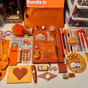 May include: A collection of orange-themed stationery and accessories including a notebook, pens, stickers, a scrunchie, a pouch, a heart-shaped notepad, a hand cream tube, and various clips arranged on a surface. The items feature various orange shades.