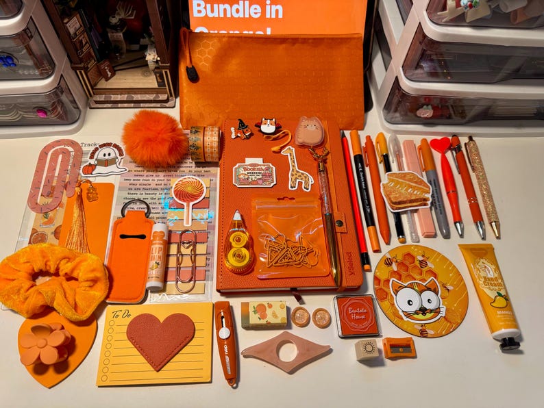May include: A collection of orange-themed stationery and accessories including a notebook, pens, stickers, a scrunchie, a pouch, a heart-shaped notepad, a hand cream tube, and various clips arranged on a surface. The items feature various orange shades.