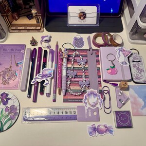 May include: A collection of purple stationery items including a notebook, pens, pencils, a 12-centimeter ruler, washi tape, stickers, a beaded lanyard with bows, a keychain, a stamp, and a circular coaster featuring a purple iris flower illustration.