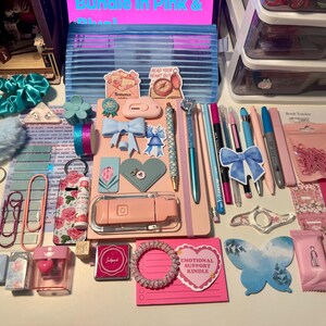 May include: A collection of stationery and accessories in pink and blue tones, including a notebook, pens, stickers, a pencil case, hair scrunchies, paper clips, a lip balm, a small ink pad, and various decorative items arranged on a flat surface.