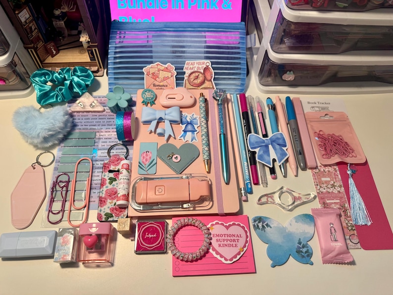May include: A collection of stationery and accessories in pink and blue tones, including a notebook, pens, stickers, a pencil case, hair scrunchies, paper clips, a lip balm, a small ink pad, and various decorative items arranged on a flat surface.