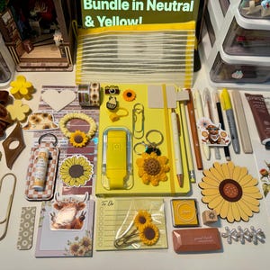 May include: A collection of yellow and neutral stationery items including a yellow notebook, pens, sunflower-themed clips, stickers, a brown velvet scrunchie, a mesh pouch, a tube of cream, and various decorative paper goods arranged on a flat surface.