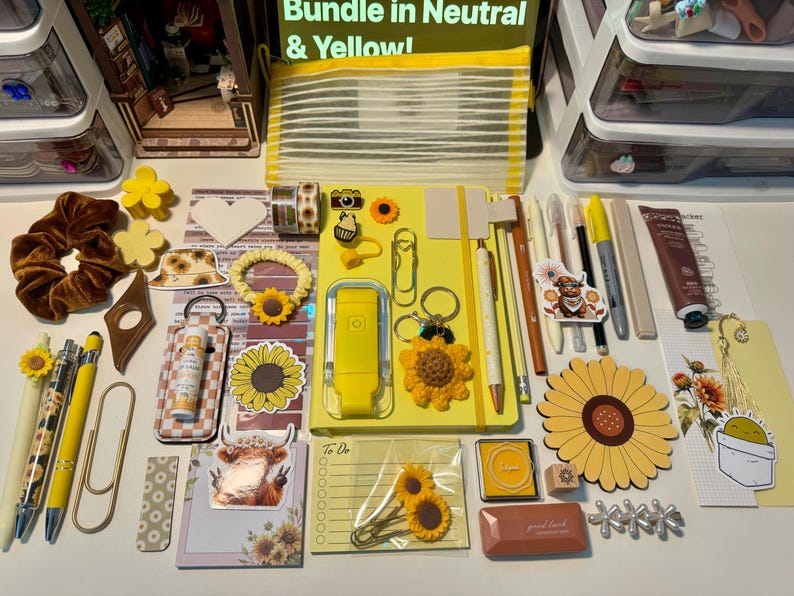 May include: A collection of yellow and neutral stationery items including a yellow notebook, pens, sunflower-themed clips, stickers, a brown velvet scrunchie, a mesh pouch, a tube of cream, and various decorative paper goods arranged on a flat surface.