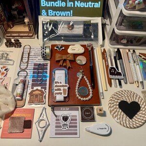 May include: A collection of stationery items including a brown notebook, pens, pencils, a pearl beaded keychain, a heart-shaped coaster, stickers, a clear pencil case, a white correction tape, a scrunchie, and various paper clips arranged on a flat surface.