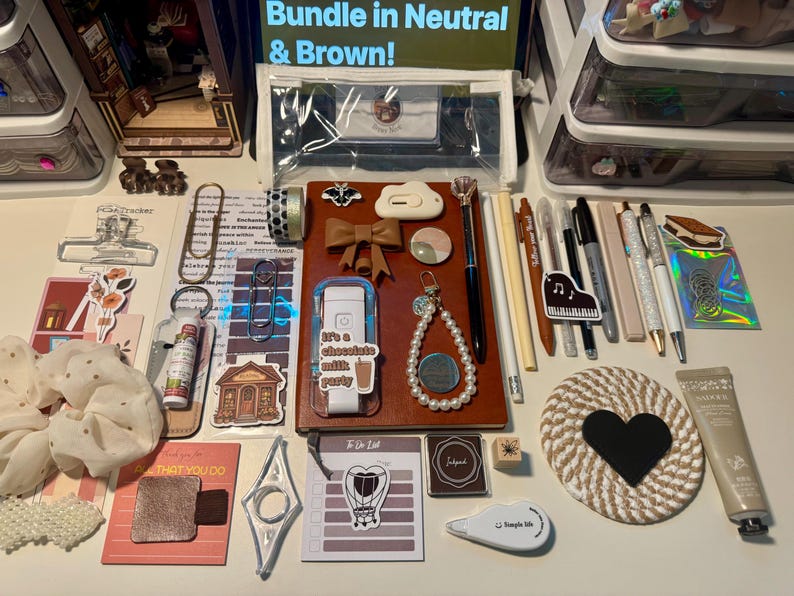 May include: A collection of stationery items including a brown notebook, pens, pencils, a pearl beaded keychain, a heart-shaped coaster, stickers, a clear pencil case, a white correction tape, a scrunchie, and various paper clips arranged on a flat surface.