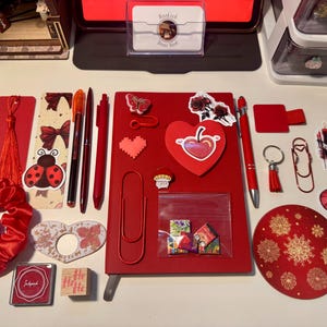 May include: A collection of red stationery items including a notebook, pens, a scrunchie, stickers, paper clips, a bookmark with a tassel, a stamp, an ink pad, and a circular coaster. The items feature various designs like a ladybug, heart, butterfly, and rose.