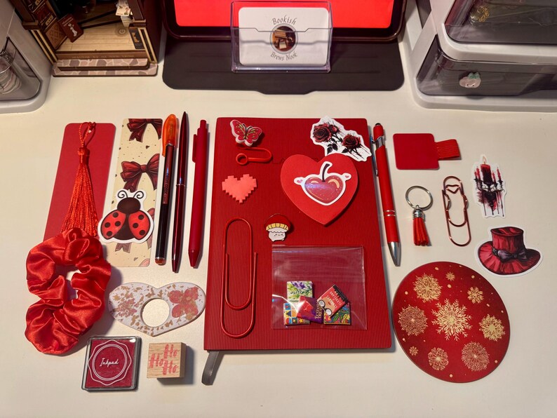 May include: A collection of red stationery items including a notebook, pens, a scrunchie, stickers, paper clips, a bookmark with a tassel, a stamp, an ink pad, and a circular coaster. The items feature various designs like a ladybug, heart, butterfly, and rose.