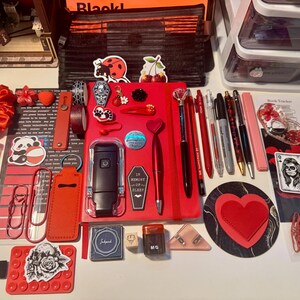May include: A collection of stationery and accessories featuring a red notebook, pens, pencils, a black book light, heart-shaped items, stickers, paper clips, a hand cream tube labeled Love, a red scrunchie, and various decorative charms arranged on a surface.