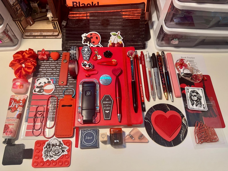 May include: A collection of stationery and accessories featuring a red notebook, pens, pencils, a black book light, heart-shaped items, stickers, paper clips, a hand cream tube labeled Love, a red scrunchie, and various decorative charms arranged on a surface.