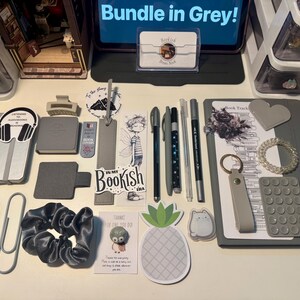 May include: A grey-themed stationery bundle featuring a notebook, pens, bookmarks, stickers, a scrunchie, a keychain, and various desk accessories. The collection includes items with text such as Bundle in Grey, Listening to Audiobooks is Reading, and In My Bookish Era, arranged on a flat surface.