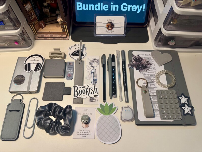 May include: A grey-themed stationery bundle featuring a notebook, pens, bookmarks, stickers, a scrunchie, a keychain, and various desk accessories. The collection includes items with text such as Bundle in Grey, Listening to Audiobooks is Reading, and In My Bookish Era, arranged on a flat surface.