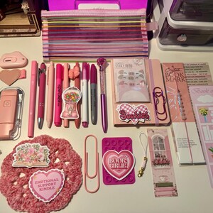 May include: A collection of pink and purple stationery items including a striped pencil case, various pens, markers, a notebook with pig-themed sticky notes, bookmarks, a crochet coaster, paper clips, and stickers with text like Bookish and Book Girlie.