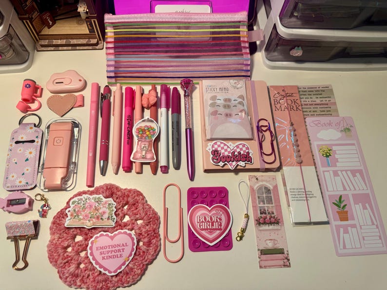 May include: A collection of pink and purple stationery items including a striped pencil case, various pens, markers, a notebook with pig-themed sticky notes, bookmarks, a crochet coaster, paper clips, and stickers with text like Bookish and Book Girlie.