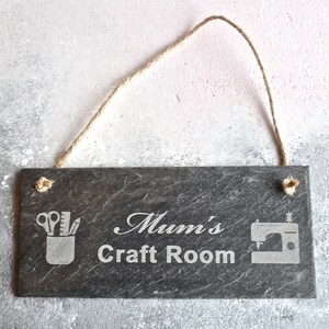 May include: Black slate sign with white text that reads "Mum's Craft Room". The sign has a rope for hanging and features a white silhouette of a sewing machine and a pencil holder with scissors, a ruler, and a pencil.