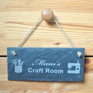 May include: A black slate sign with white text that reads "Mum's Craft Room". The sign has a rope hanger and a wooden knob. The sign also has two icons, one of scissors and pencils in a cup and the other of a sewing machine.