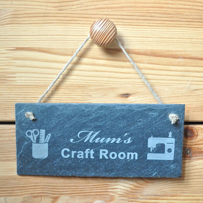 May include: A black slate sign with white text that reads "Mum's Craft Room". The sign has a rope hanger and a wooden knob. The sign also has two icons, one of scissors and pencils in a cup and the other of a sewing machine.