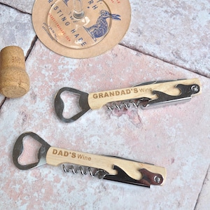 May include: Two wooden wine bottle openers with metal bottle openers and corkscrews. One opener says "Grandad's Wine" and the other says "Dad's Wine".