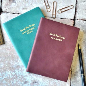 May include: Two hardcover notebooks, one teal and one burgundy, with gold lettering that reads "Sweet Pea Design APPOINTMENT BOOK" and "Sweet Pea Design PLANNER." Gold paperclips and a black pen with gold accents are also present.