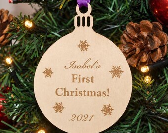 May include: A round, light-colored wooden ornament featuring a purple ribbon loop at the top. The text "Isobel's First Christmas!" and "2021" are engraved in the center, surrounded by five small, etched snowflake designs on the surface.