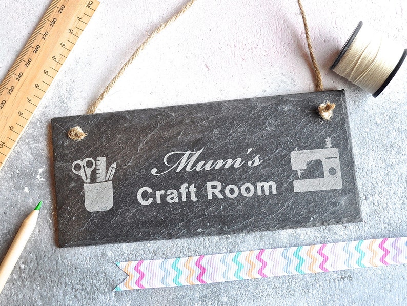 May include: Black slate sign with the text "Mum's Craft Room" engraved in white. The sign also features a silhouette of a sewing machine and a pencil holder with scissors, a ruler, and pencils.