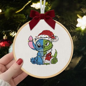 May include: Embroidered Christmas ornament featuring a split design of Stitch and the Grinch, both in Santa hats and festive lights. The ornament is round, framed in wood, and has a red velvet bow at the top for hanging.