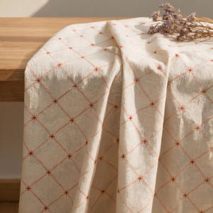 May include: A cream-coloured fabric runner featuring a repeating diamond grid pattern in a rust-orange hue. Each intersection of the grid is marked by a small, four-pointed star design. The textile has a visible weave texture and soft folds.