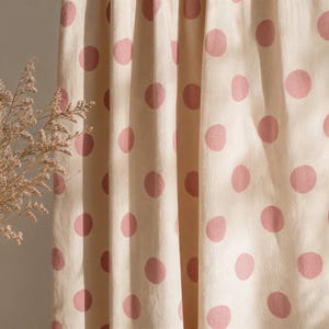 May include: A cream-coloured fabric curtain featuring a repeating pattern of soft pink polka dots. The material has visible vertical folds and a textured weave, with a small cluster of dried, light brown botanical stems visible on the left side.