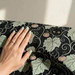 May include: A black fabric featuring a repeating pattern of light green pumpkin leaves, small pale pink pumpkins, and stylized black cat faces with bright yellow eyes. The design includes swirling vine details across the dark background.