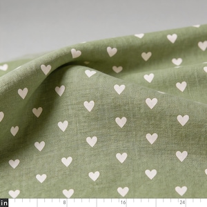 Puede incluir: Tela de color verde salvia con un patrón repetitivo de pequeños corazones blancos. En el borde inferior se observa una cinta métrica blanca con medidas en pulgadas de 0 a 24, que indica la escala del estampado de corazones en el tejido.