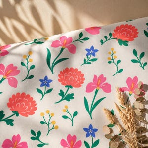 May include: A white fabric featuring a repeating pattern of pink irises, coral-coloured chrysanthemums, small blue five-petalled flowers, and yellow berry-like sprigs with green leaves, accompanied by dried wheat stalks in the corner.