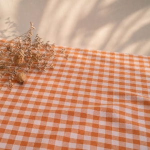 Op de afbeelding: Een textielstof met een klassiek oranje en wit vichy-ruitpatroon. Op het oppervlak van de gevouwen stof ligt een klein, delicaat arrangement van gedroogde, lichtbruine wilde bloemen met dunne stengels en zaaddoosjes.