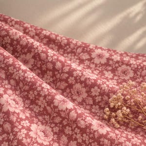 May include: A folded fabric featuring a repeating floral pattern in shades of dusty rose and light pink. The textile displays intricate illustrations of various blossoms and leaves across the surface, accompanied by a small sprig of dried flowers.