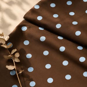 May include: A dark brown fabric featuring a repeating pattern of light blue polka dots. The textile is folded, creating soft drapes and shadows across the surface. A single dried eucalyptus stem with small, rounded leaves rests on the fabric.