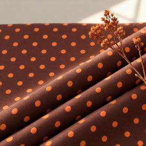 May include: A dark brown fabric featuring a repeating pattern of small, evenly spaced orange polka dots. The material is draped in soft folds, and a dried, light brown botanical stem rests across the surface of the textile.