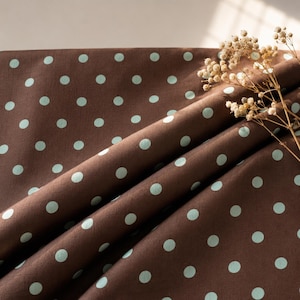 May include: A dark brown fabric featuring a repeating pattern of small, light mint green polka dots. The material is draped in soft folds, creating natural shadows and highlights across the surface, with a sprig of dried baby's breath flowers.