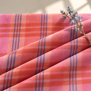Op de afbeelding: Een gevouwen stuk stof met een ruitpatroon in tinten roze, oranje en blauw. Het textiel vertoont een raster van verticale en horizontale strepen. Een klein takje met blauwe bloemen rust op de rechterbovenhoek van de stof.
