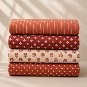May include: A stack of four fabric bolts featuring warm colour palettes. The top bolt has vertical white stripes on a rust background, the second has small white floral patterns on dark red, the third has orange tree motifs on cream, and the bottom has white polka dots on rust.