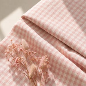 Può includere: Un tessuto a quadretti vichy rosa chiaro e bianco con una piccola e delicata composizione di fiori secchi rosa e steli di erba coda di lepre soffice appoggiati sulla superficie tessile piegata, che mostra una trama e un motivo intrecciati.