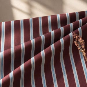 May include: A piece of fabric featuring a deep burgundy background with vertical stripes in light blue and white. The material is draped to create soft folds, and a small sprig of dried lavender is placed on the right side of the textile.