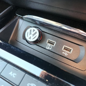 Op de afbeelding: Een close-up van een auto-console met een USB-poort en een sigarettenaansteker met het Volkswagen-logo. De aansteker heeft een zwart en zilver ontwerp met een rode accentkleur. De console is zwart met zilveren afwerking.